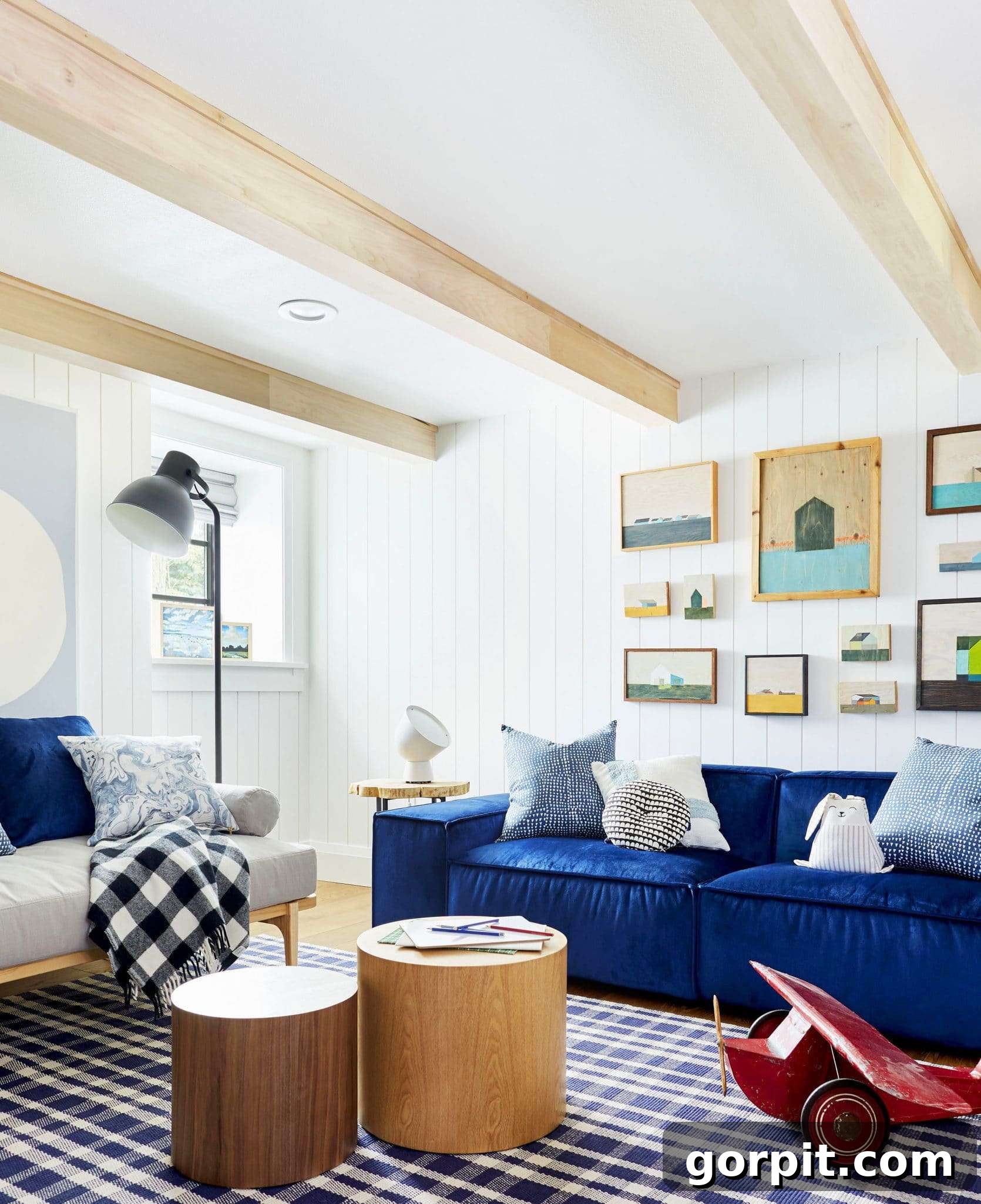 Light wood beams make an interesting ceiling for a white wood paneled room with deep blue couches. A grey futon, round wood coffee tables and part of a large wooden toy plane complete this playroom idea. 