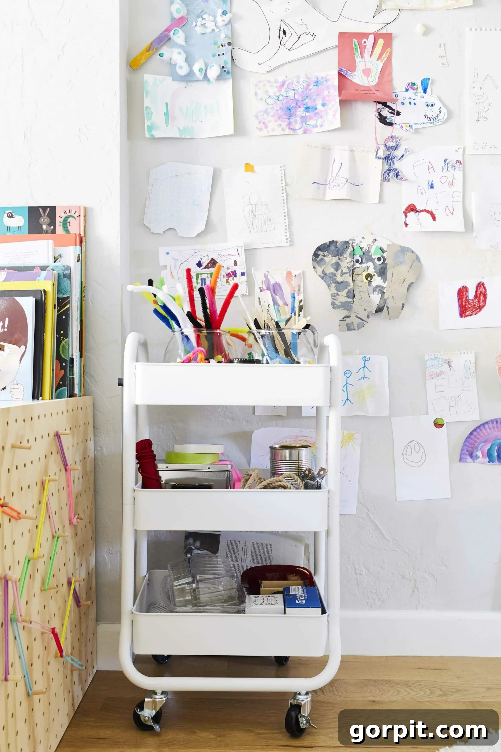 One of the best playroom ideas is to use children's artwork as decoration. A wall of kid's art includes an elephant, handprints, and more crafts. Books and an art cart are in front. 