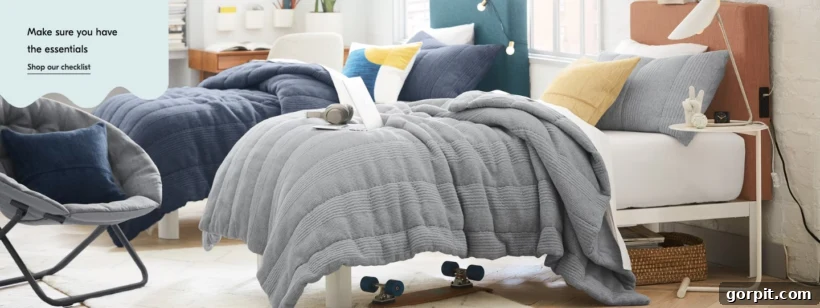 West Elm dorm room ideas include these twin beds, blue-toned bedding, a gray bucket chair, and sparse nightstands. The text in the corner reads, "Make sure you have the essentials, shop our checklist."