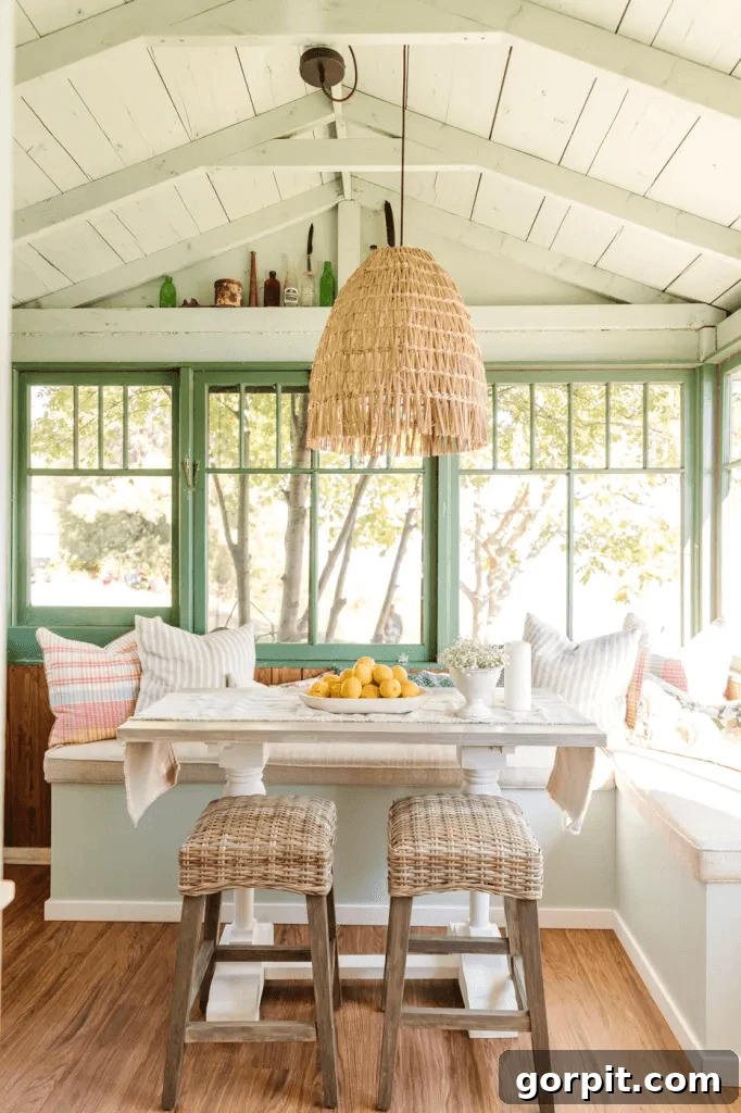 Screened Porch Dreams: Craft Your Perfect Outdoor Escape 6 This screened-in porch idea includes an eating area with built in bench seats, green windows, a white wooden table with stools and a hanging grass chandelier over the bowl of oranges.