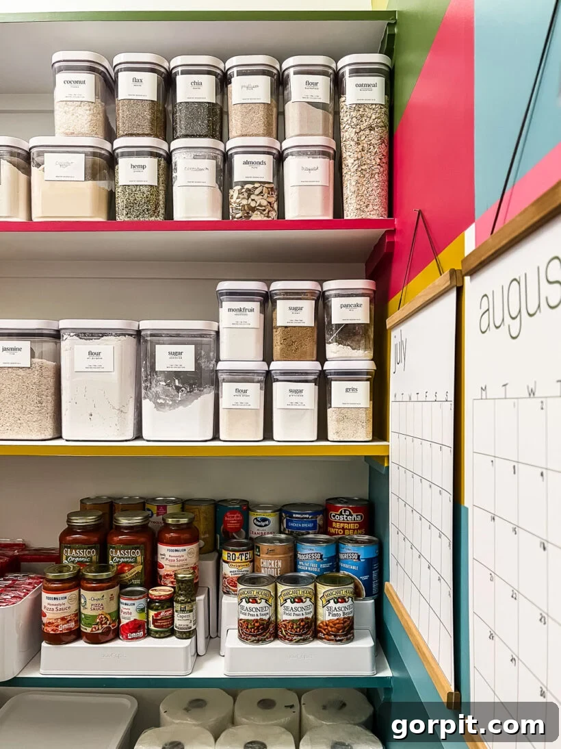 Organized canned goods on tiered risers and neatly decanted dry goods in OXO Pop containers, within a colorful pantry.