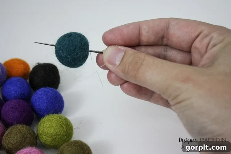 A hand holds a needle that is piercing a deep blue felted ball used to make DIY car air fresheners. 