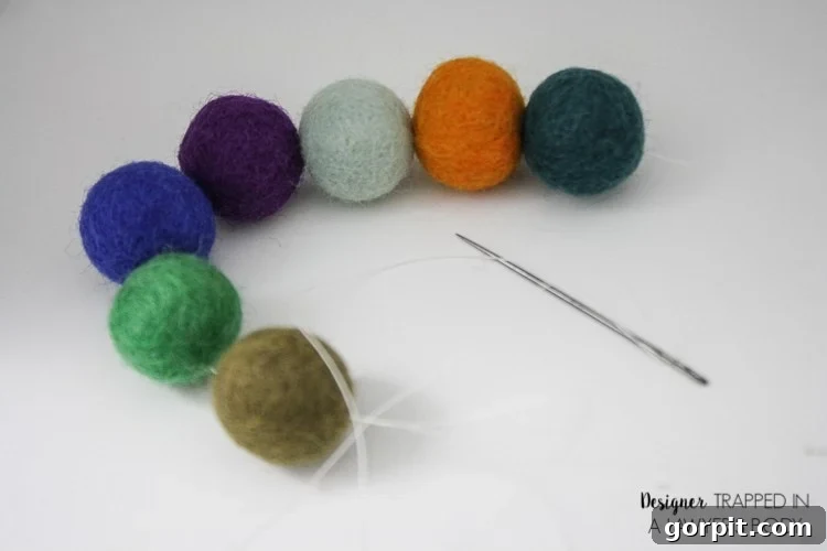 A close-up of a string of colorful small felt balls and a needle with white thread. The corner text reads, 