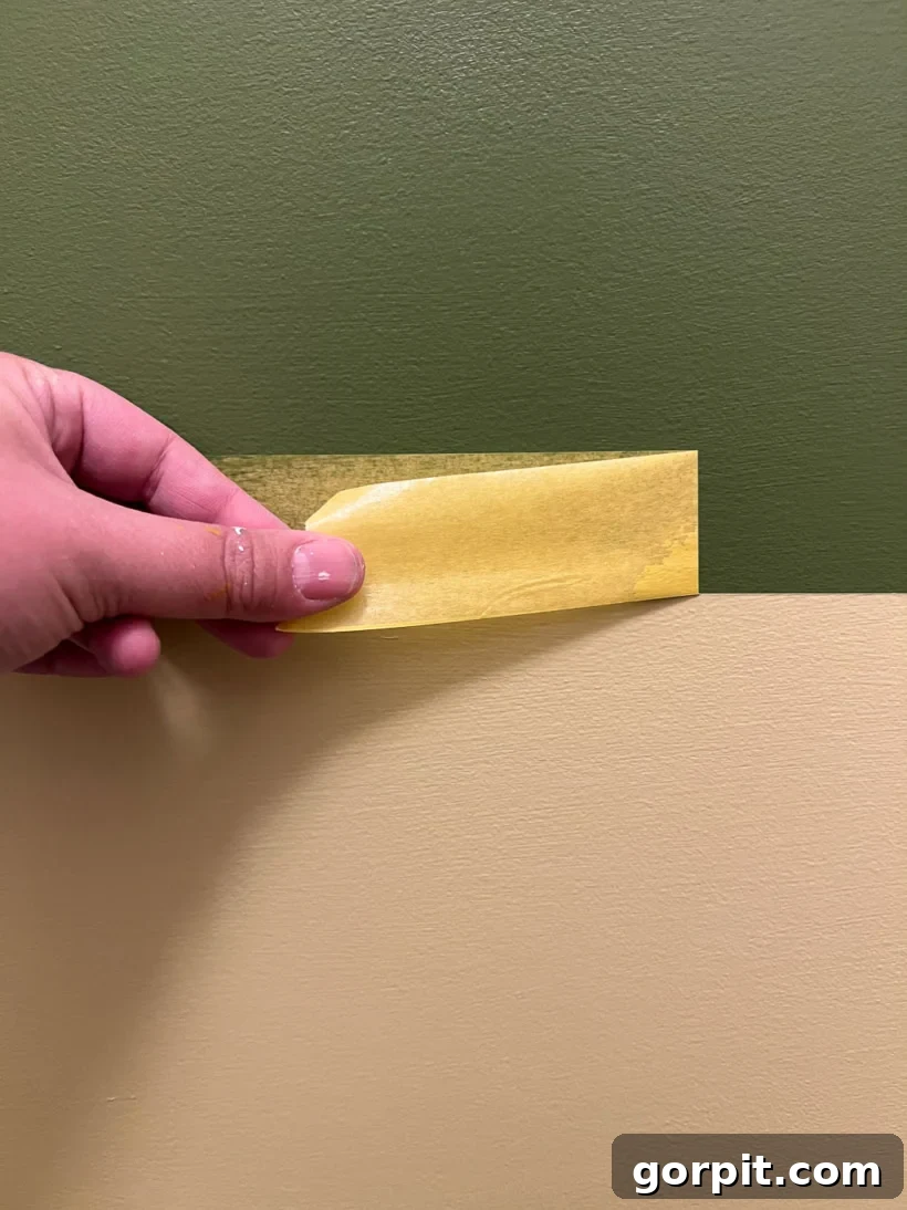Photo showing a hand carefully peeling off yellow painter's tape from a wall, revealing a clean, sharp line where two different green paint colors meet, emphasizing the importance of correct tape removal timing for flawless results.