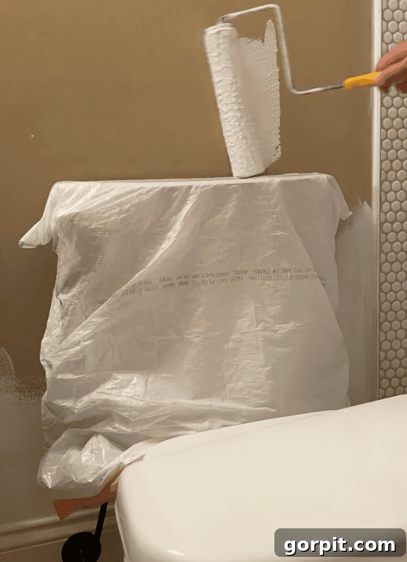 Image of a paint roller carefully painting near a toilet, with the toilet tank neatly protected by a plastic bag, demonstrating a clever hack for achieving a clean paint job in challenging bathroom spaces.
