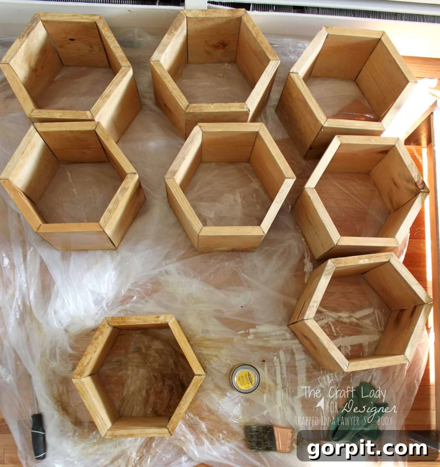 Honeycomb Shoe Rack DIY 15 Hexagon-shaped wooden shoe cubbies laid out on a plastic-covered hard wood floor, ready for staining or sealing, with a brush, screwdriver, and wood finish can nearby. Text in the lower right corner reads,